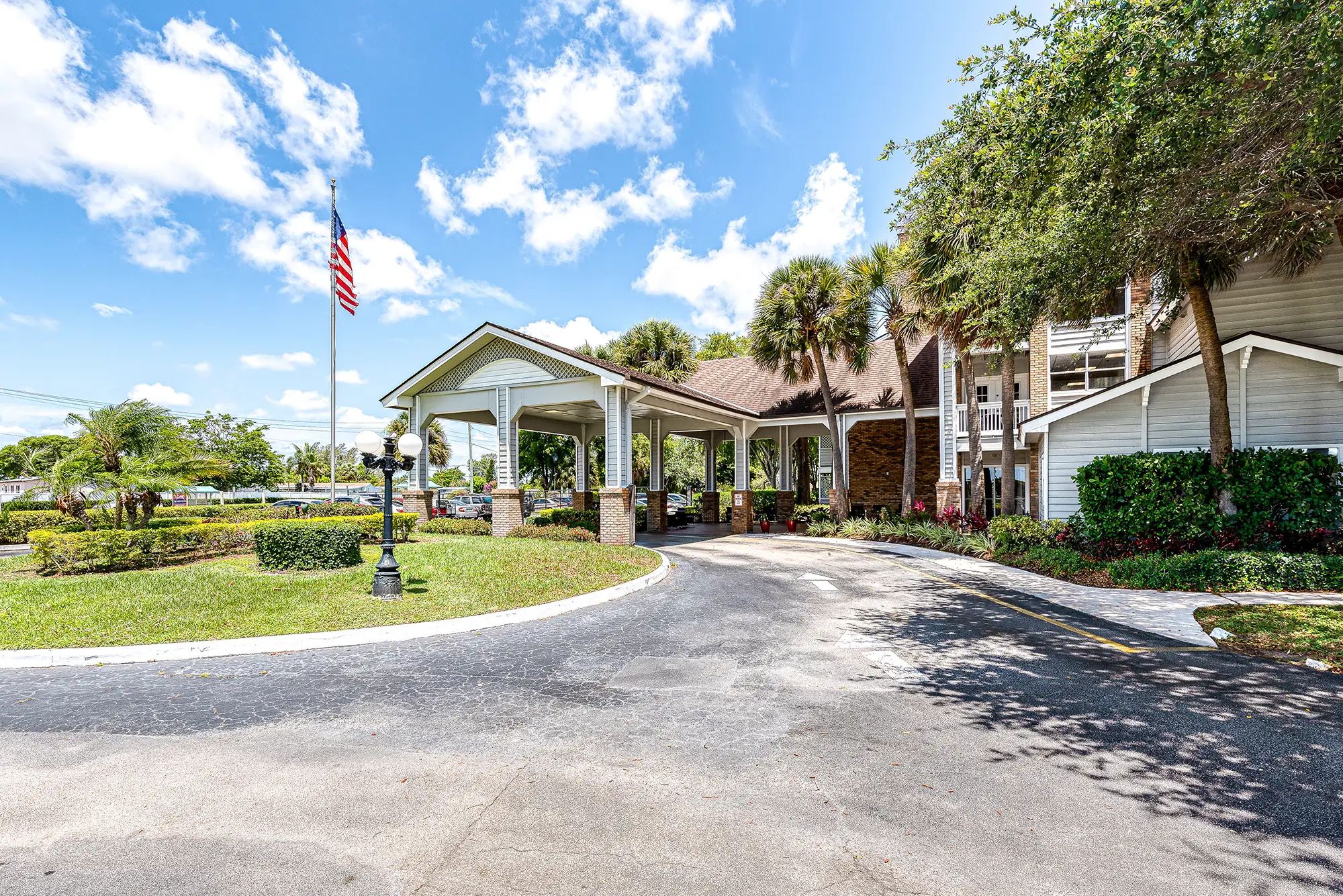 An outside view of the entrance at The Meridian at Lantana in FL. The entrance has a large round about with lots of trees surrounding the property.