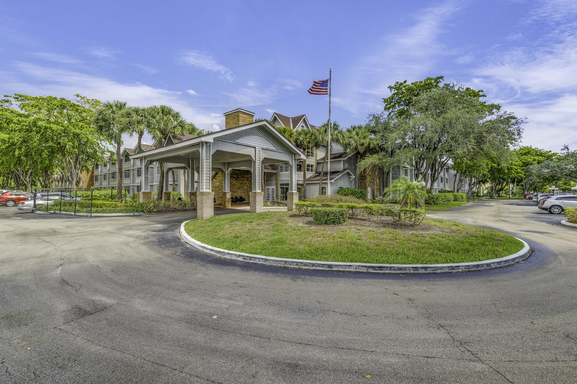 An outside view of the entrance at The Meridian at Lantana in FL. The entrance has a large round about with lots of trees surrounding the property.